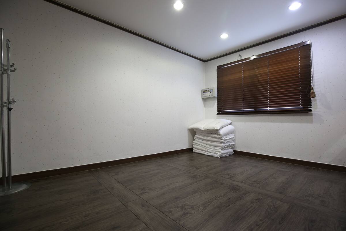 A room with a wooden floor, a window with blinds, a radiator, and a stack of white towels.