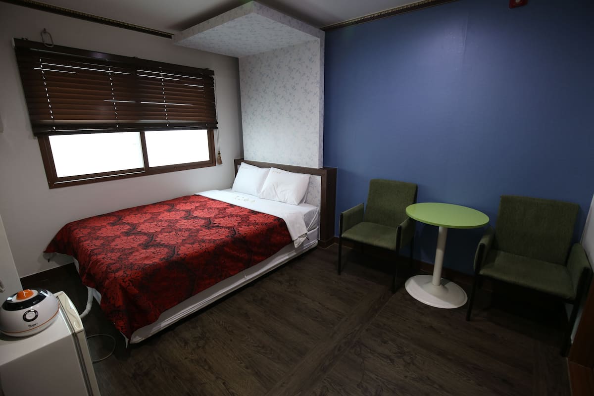 A room with a bed, two chairs, a small table, and a window with blinds.