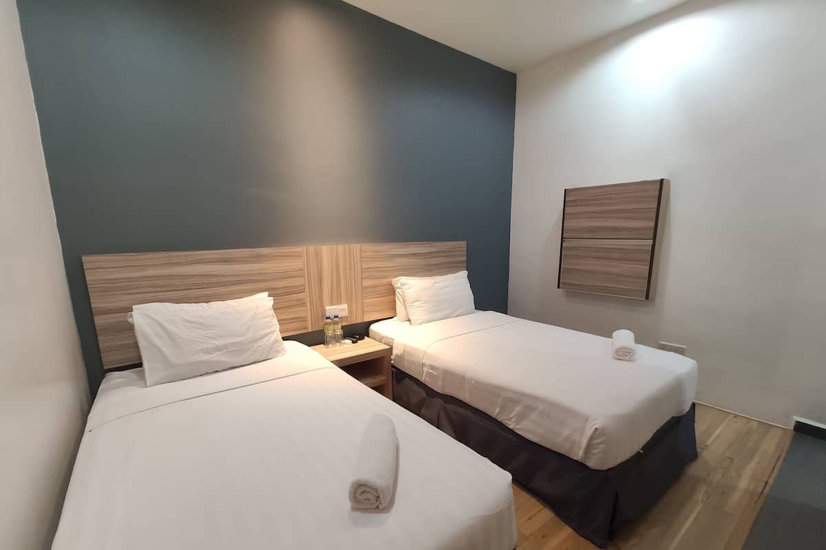 A hotel room with two beds, each with white linens and a rolled towel. There is a wooden headboard on the left bed, and a wall-mounted shelf with a lamp and a small table.