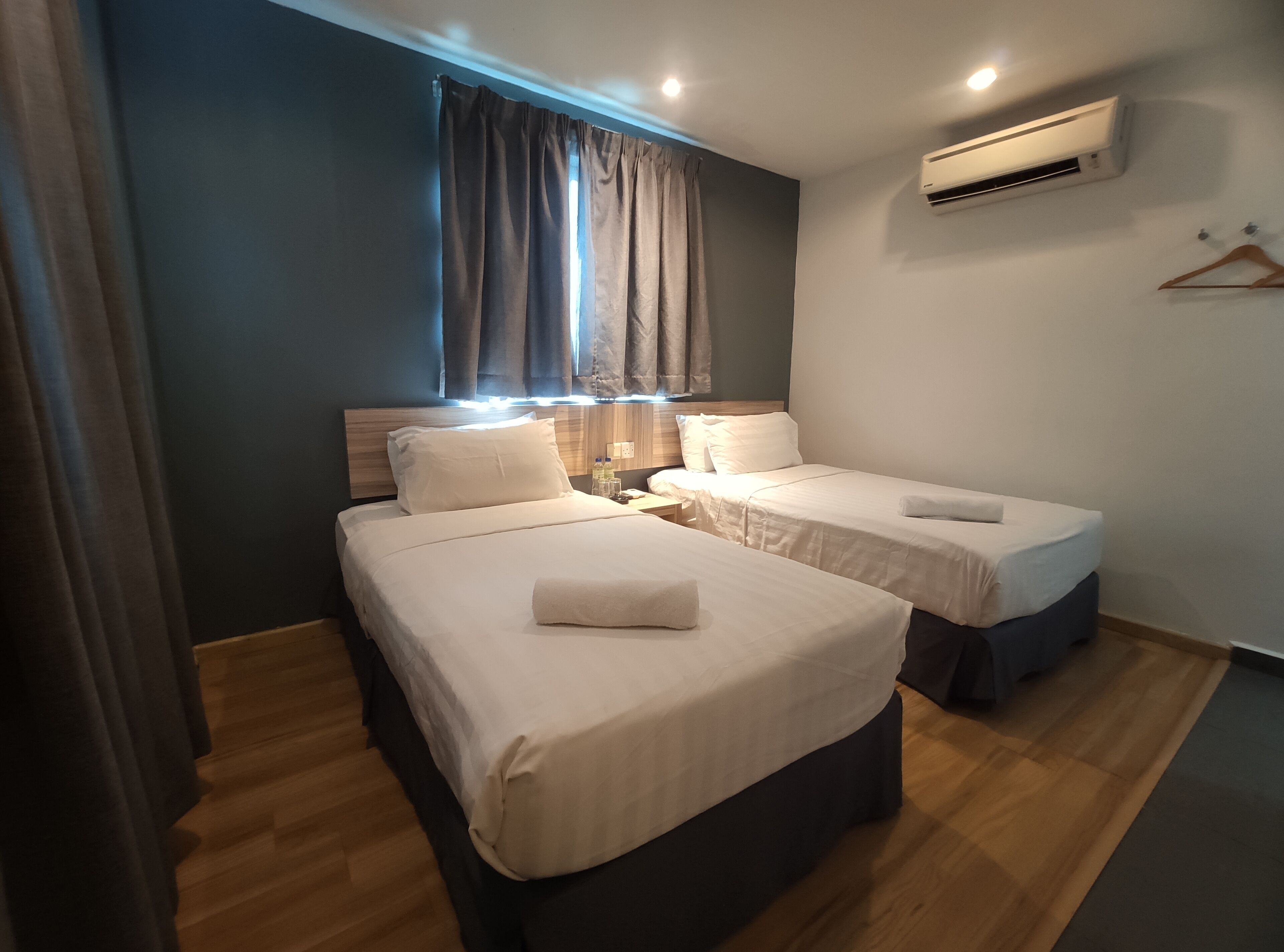 A hotel room with two beds, a window with curtains, an air conditioning unit, and a wooden floor.