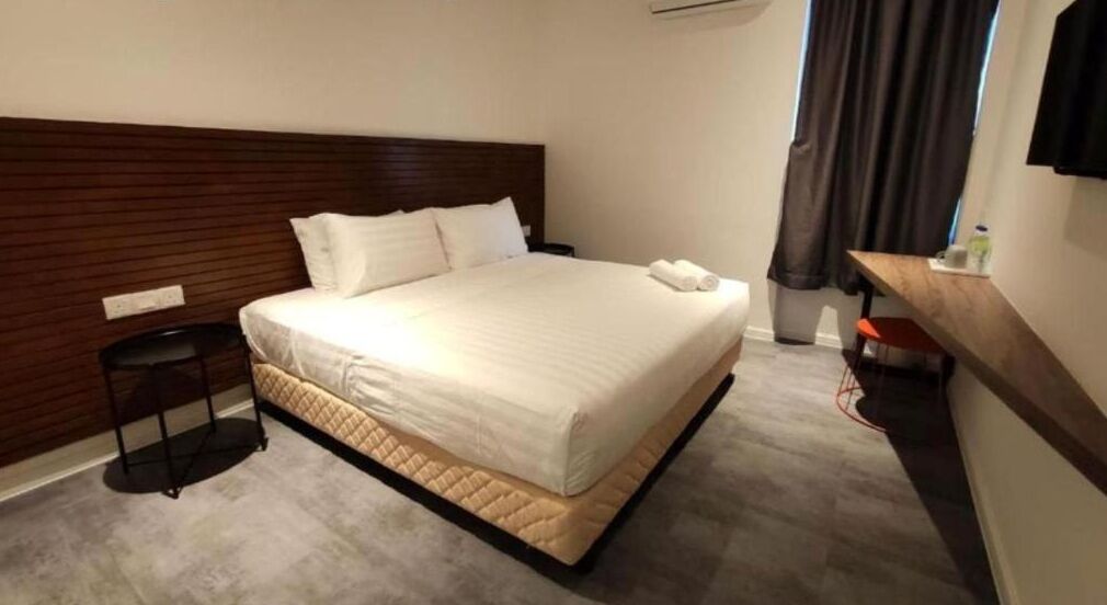 A hotel room with a large bed, a wooden headboard, a bedside table with a lamp, and a small stool.