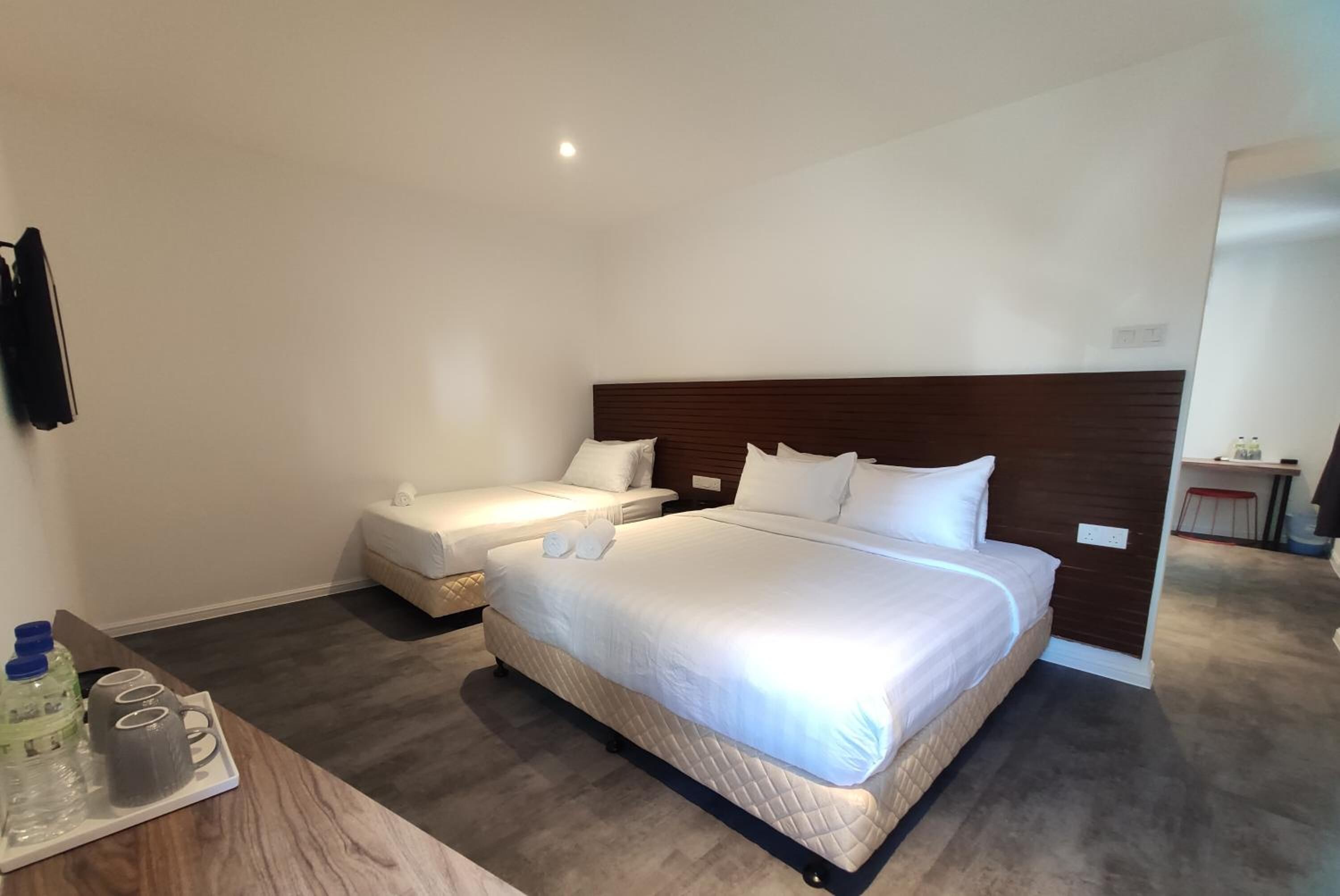 Una camera d'albergo con un letto grande, un letto più piccolo, un comodino con una bottiglia e dei bicchieri, e uno specchio a parete.