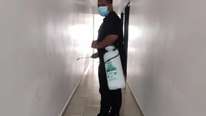Una persona parada en un pasillo con piso de baldosas y paredes blancas, sosteniendo un atomizador y un recipiente blanco con etiqueta.
