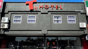 Un hotel con un logotipo destacado con la letra 'T' y señalización que dice 'HOTEL'. Se observan unidades de aire acondicionado en la fachada, y ventanas con cortinas.