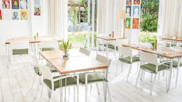Een lichte eetruimte met witte stoelen en tafels, omgeven door grote ramen en kunstwerken aan de muren.