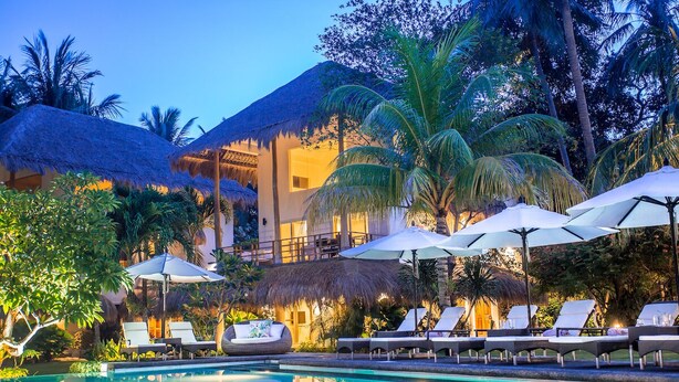 A poolside area with lounge chairs and umbrellas, surrounded by palm trees and a building with a thatched roof.