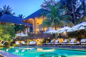 Un espace aménagé au bord de la piscine, avec des chaises longues et des parasols, entouré de palmiers et d’un bâtiment doté d’un toit de chaume.