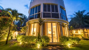 A modern, multi-story building with large windows and a circular design, surrounded by lush greenery and palm trees.