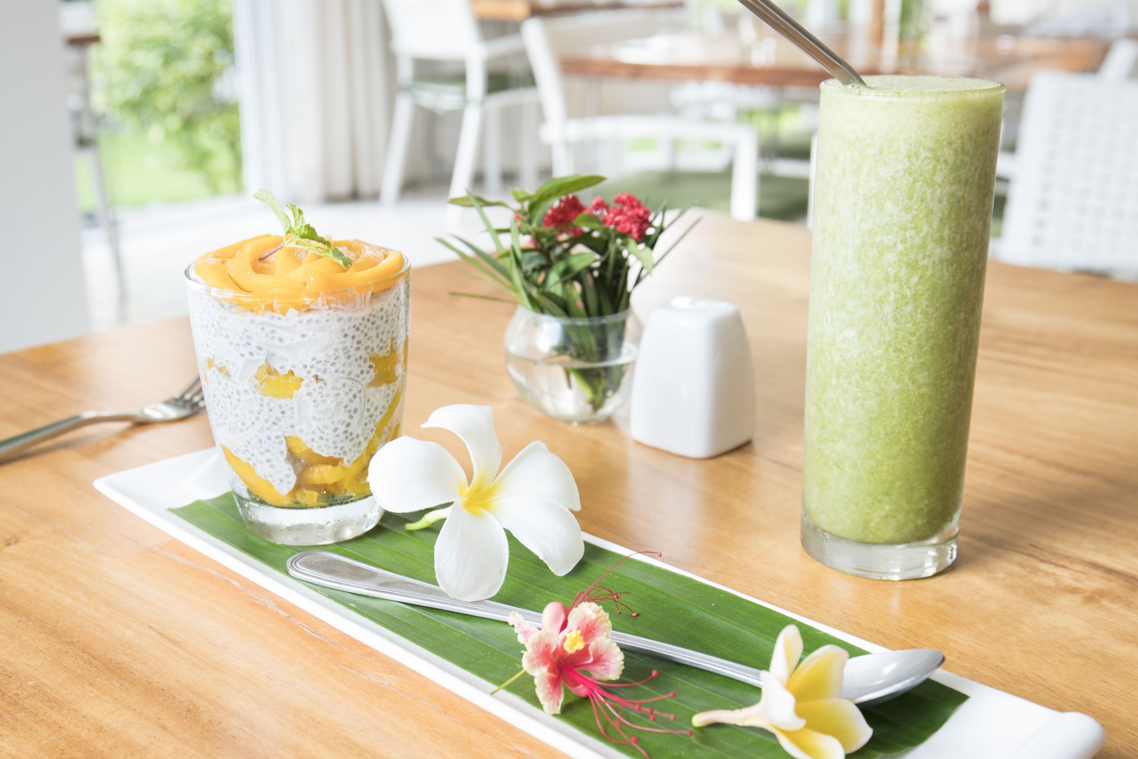 Een tafel met een glas mangosmoothie, een groene smoothie en een toetje met mango op een bananenblad, inclusief bloemen en een lepel.