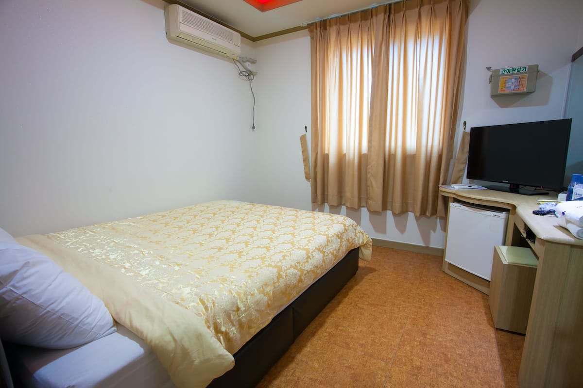A hotel room with a bed, a desk with a television, a small refrigerator, and an air conditioning unit.