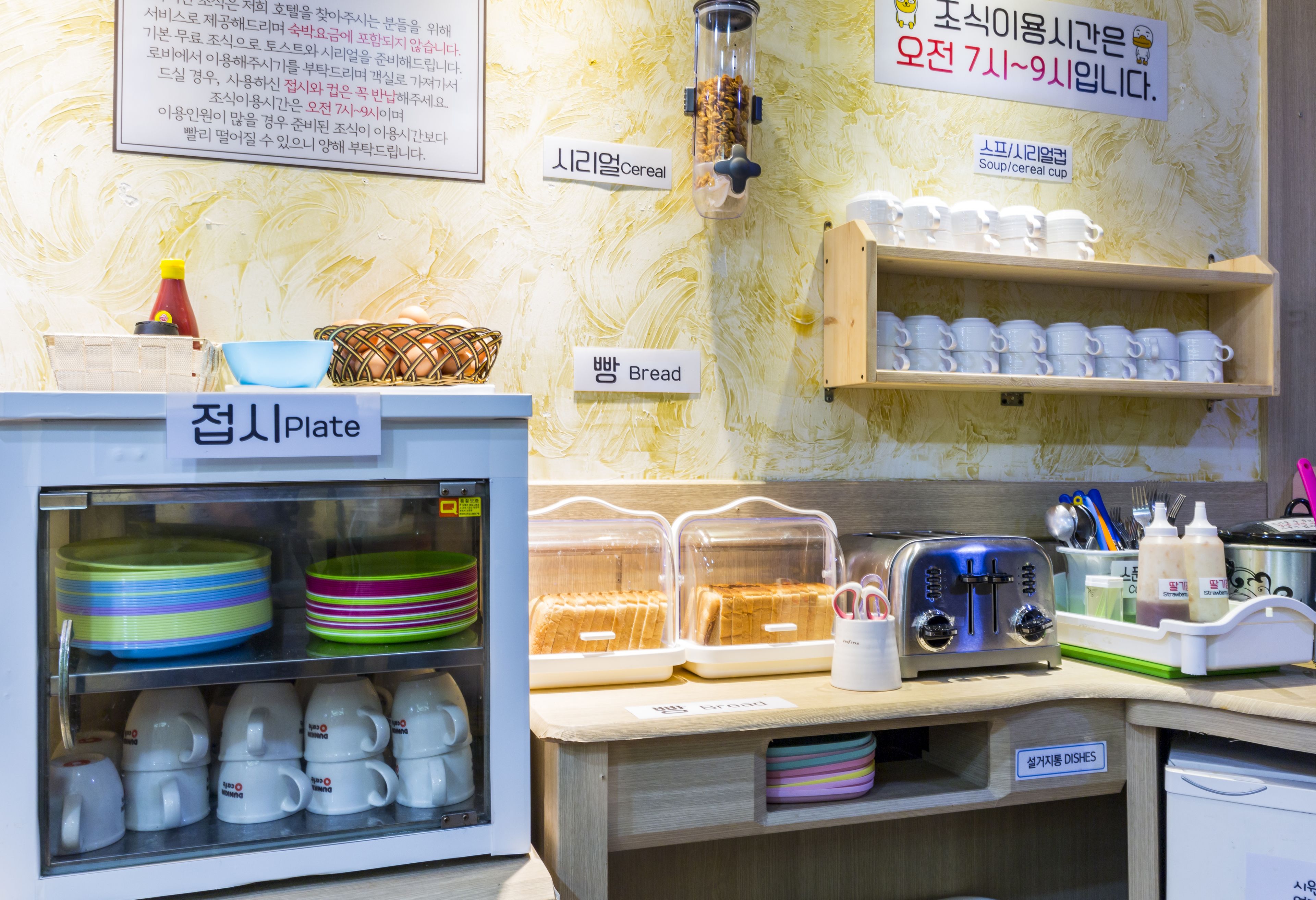 A kitchen area with labeled storage for bread, cereal, and soup. There are also plates, cups, and a toaster on the counter.