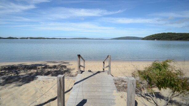 Un sendero de madera que conduce a una playa arenosa con aguas tranquilas y colinas al fondo.