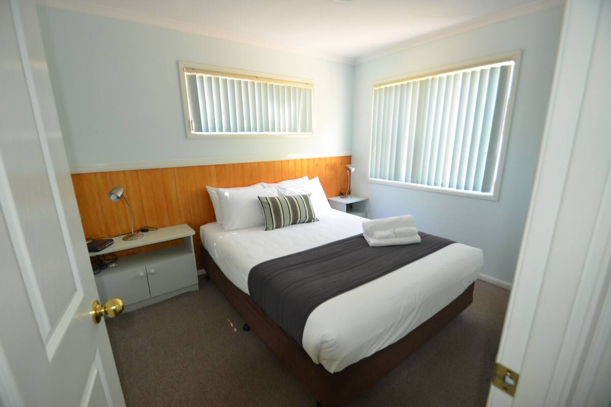 A hotel room with a bed, bedside tables, a nightstand, and a window with blinds.