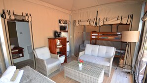 Una habitación pequeña y funcional con sofá, una mesita, una silla, una estantería, una cama litera y una lámpara.
