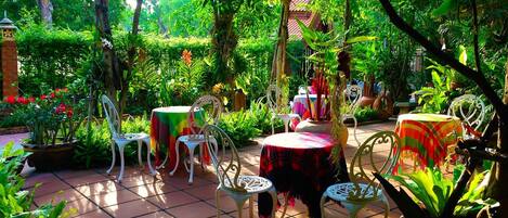 Un patio al aire libre con suelo de baldosas, sillas de metal blanco y mesas redondas cubiertas con manteles de colores.