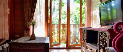 A room with a bed, a television, a table with a plant, and a sliding glass door leading to a balcony with greenery.