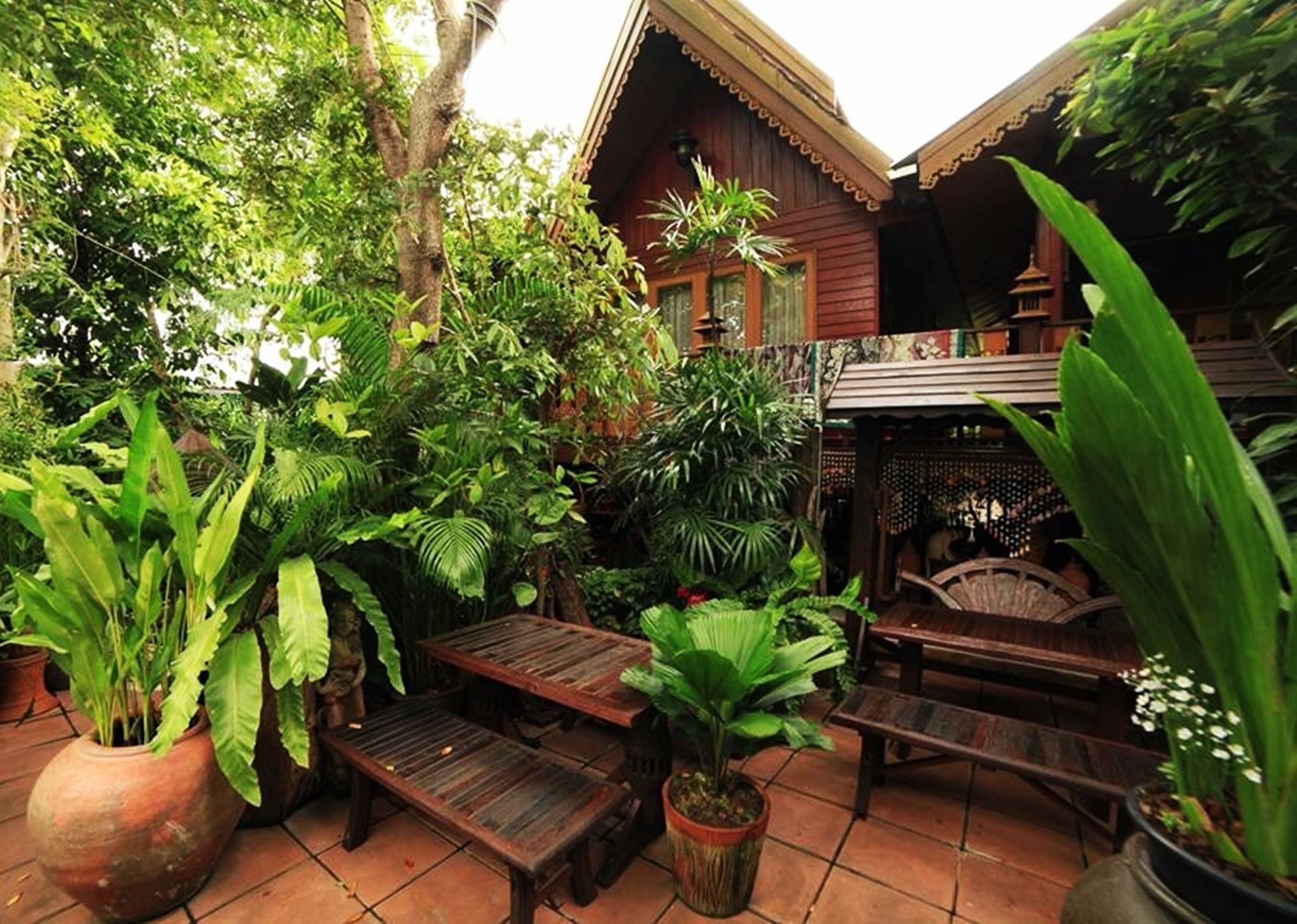 A wooden outdoor seating area with benches and tables, surrounded by lush greenery and potted plants.