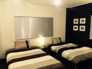 Habitación de hotel con dos camas, una ventana con persianas, una mesita de noche con lámpara y una pared decorada con cuadros.