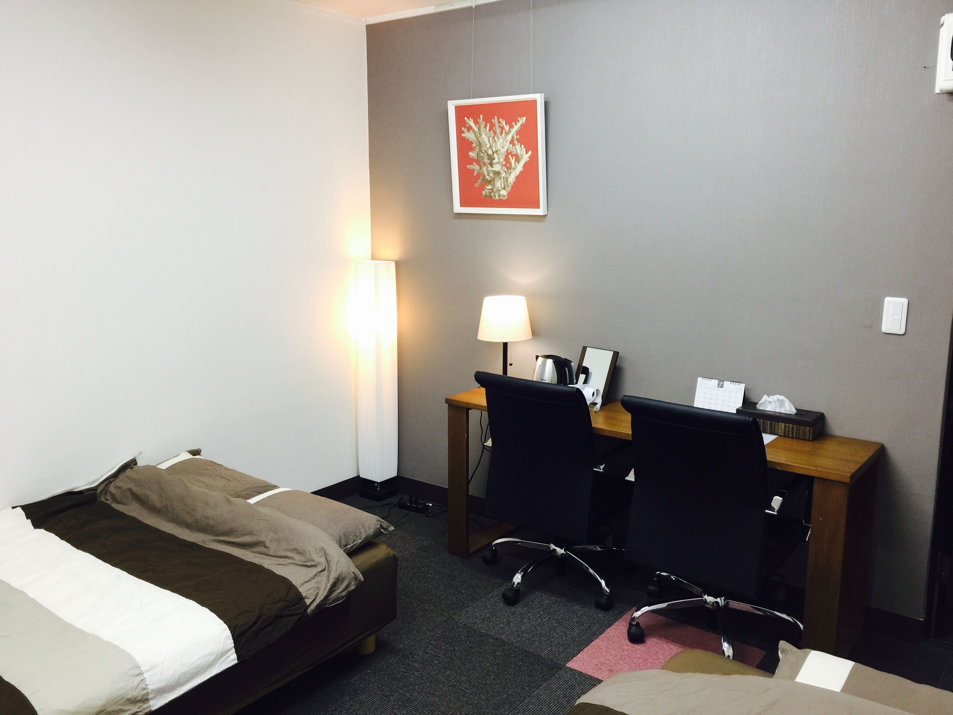 A hotel room with a bed, a desk with a chair, a lamp, a picture on the wall, and a calendar on the desk.