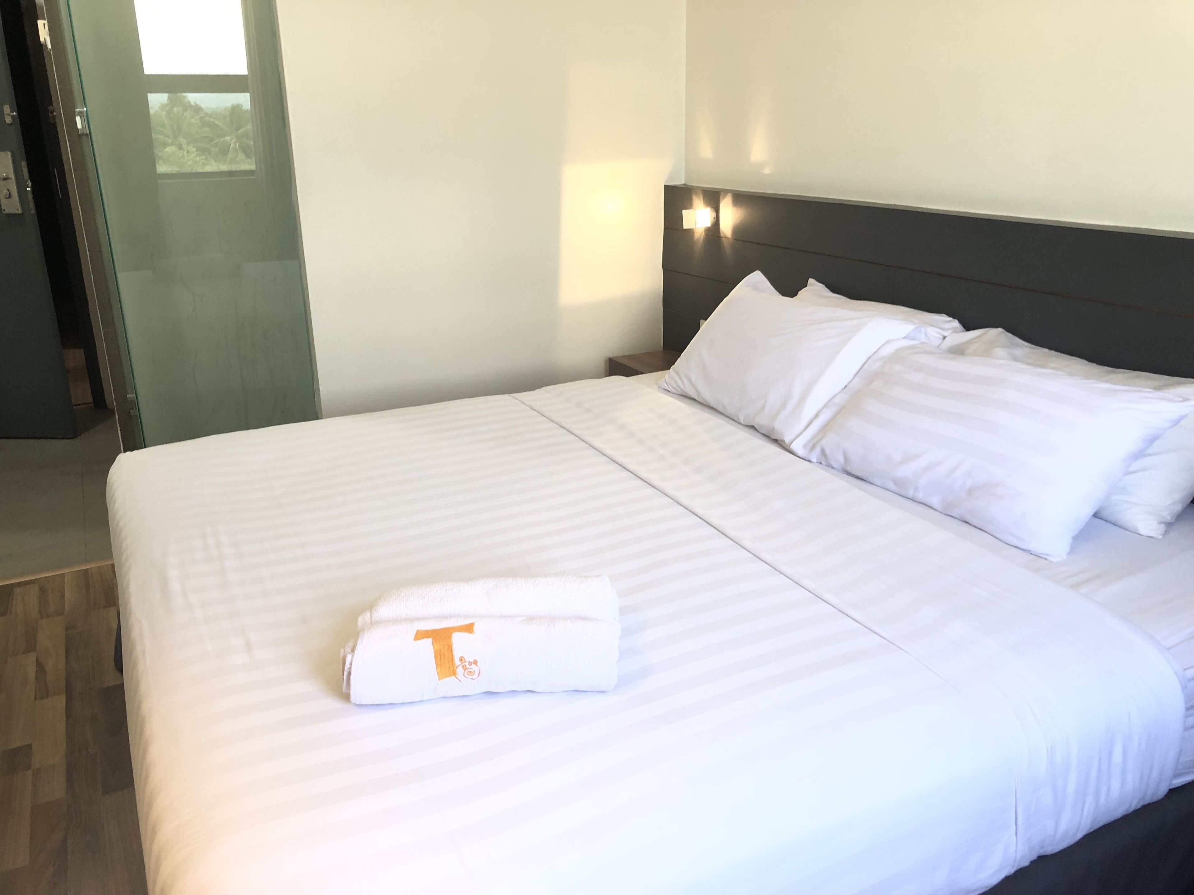 Una cama bien hecha con sábanas y almohadones blancos, una toalla doblada con un logotipo y una mesita de noche con una lámpara.