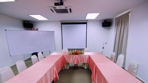 Un salón de conferencias con una larga mesa cubierta con un mantel rosa, sillas blancas, una pizarra, una pantalla de proyector y un ramo de flores sobre la mesa.