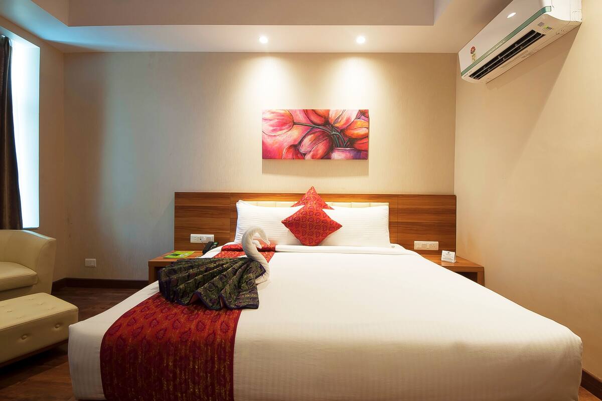 A hotel room with a neatly made bed, a wooden headboard, a painting on the wall, and a window with curtains.