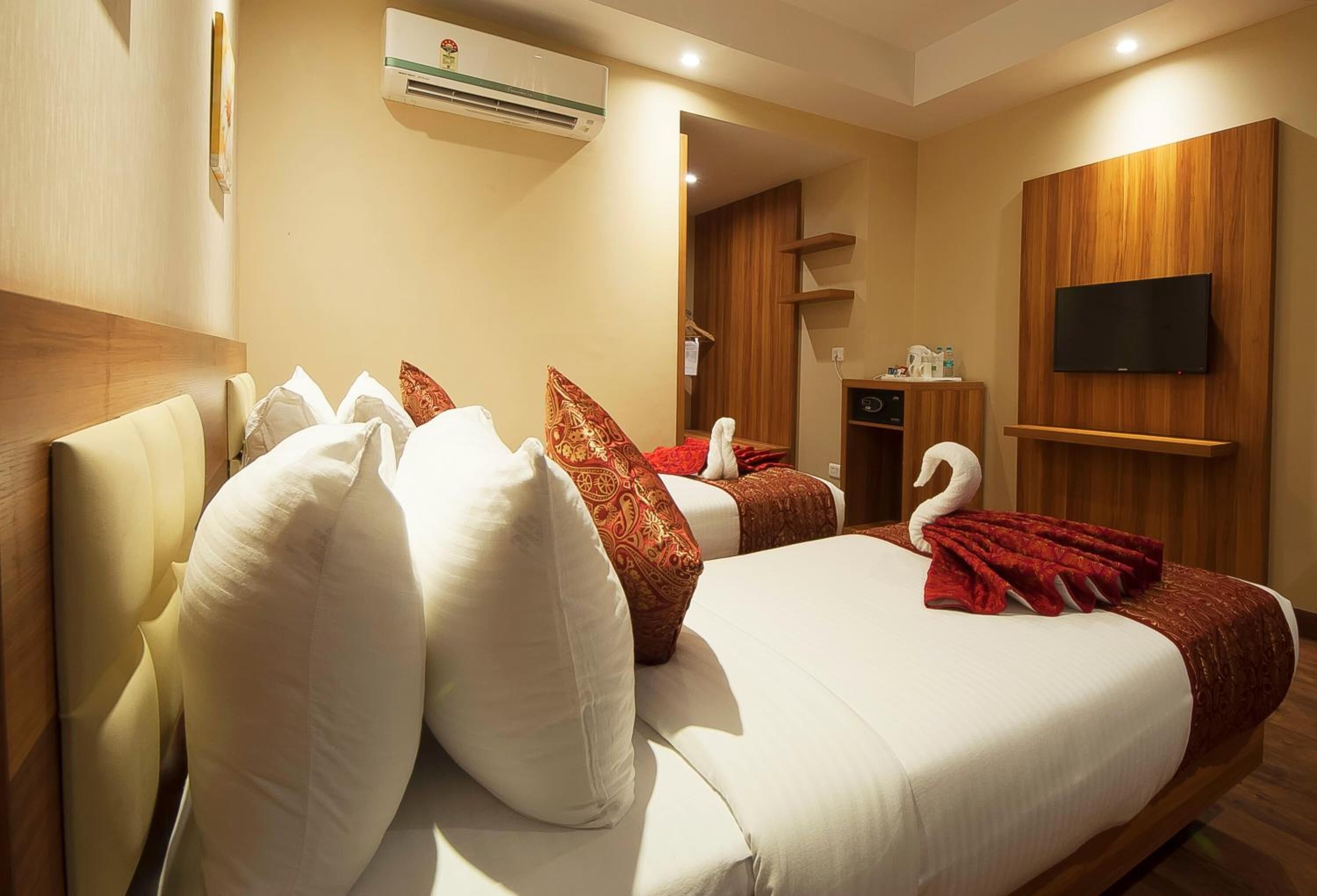 Habitación de hotel con dos camas, cada una con sábanas blancas y almohadas decorativas. Hay un televisor de pantalla plana montado en la pared, un equipo de aire acondicionado sobre las camas y un mueble de madera con un televisor.