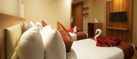 Habitación de hotel con dos camas, cada una con sábanas blancas y almohadas decorativas. Hay un televisor de pantalla plana montado en la pared, un equipo de aire acondicionado sobre las camas y un mueble de madera con un televisor.