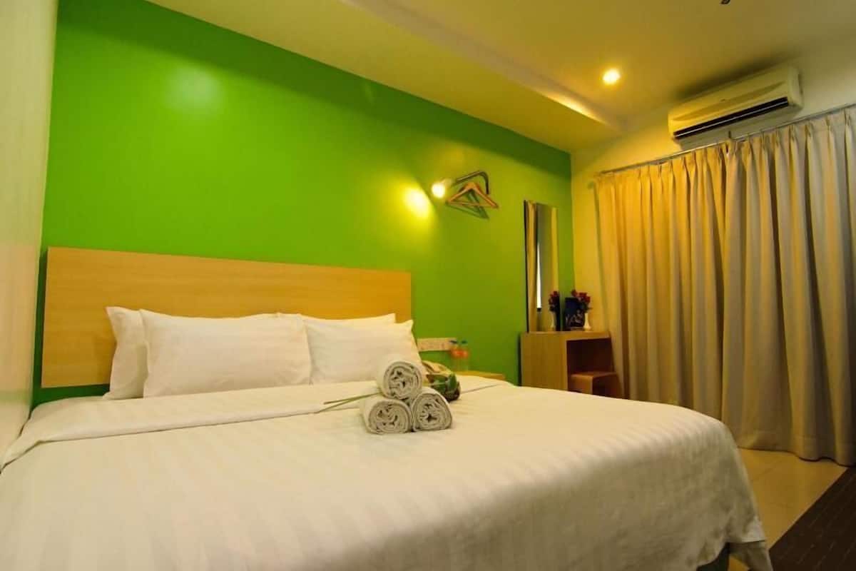 A hotel room with a green wall, a wooden headboard, a bed with white linens, a rolled towel, a nightstand with a lamp, and a window with curtains.