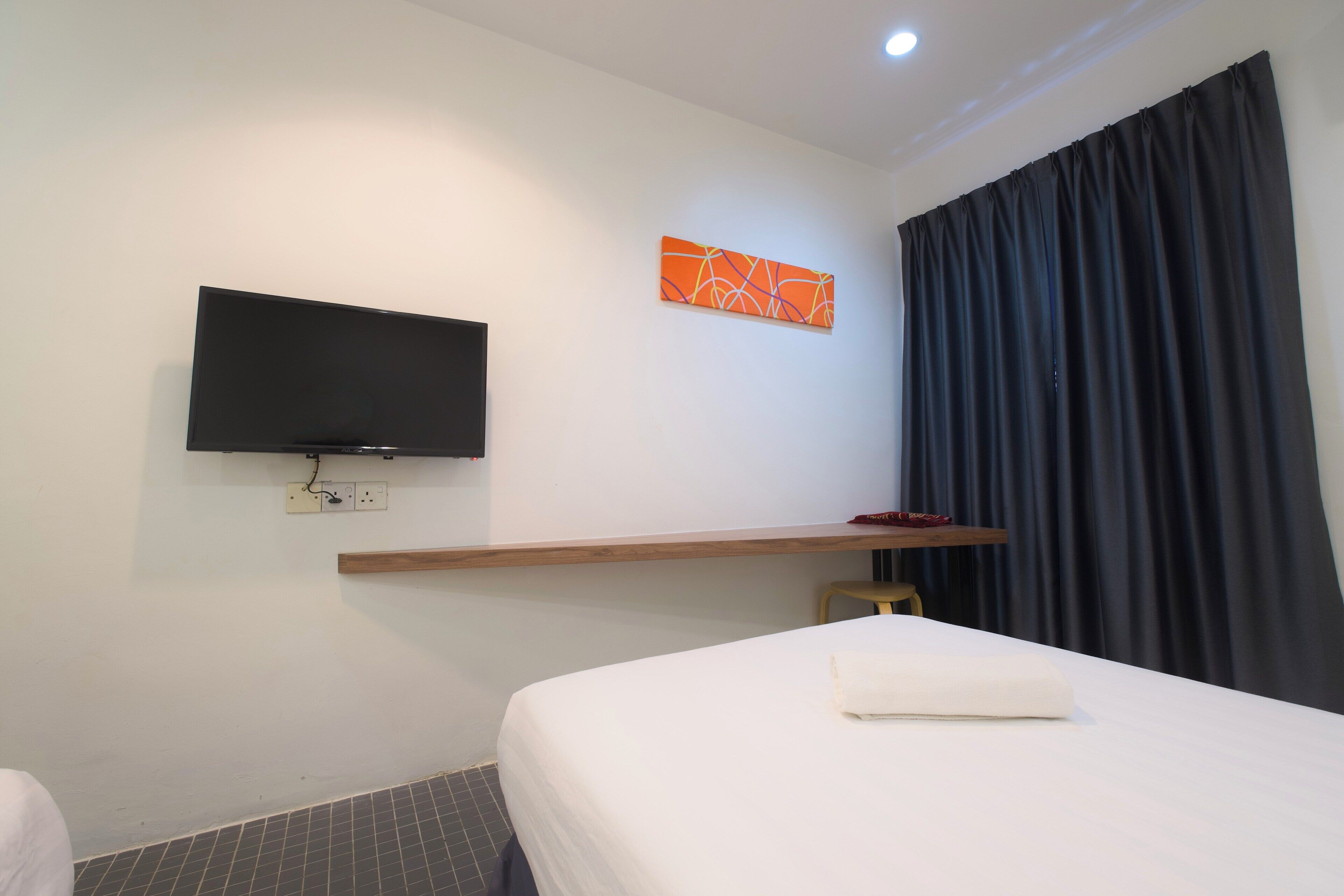 A hotel room with a bed, a TV mounted on the wall, a small wooden shelf, and a window with dark curtains.