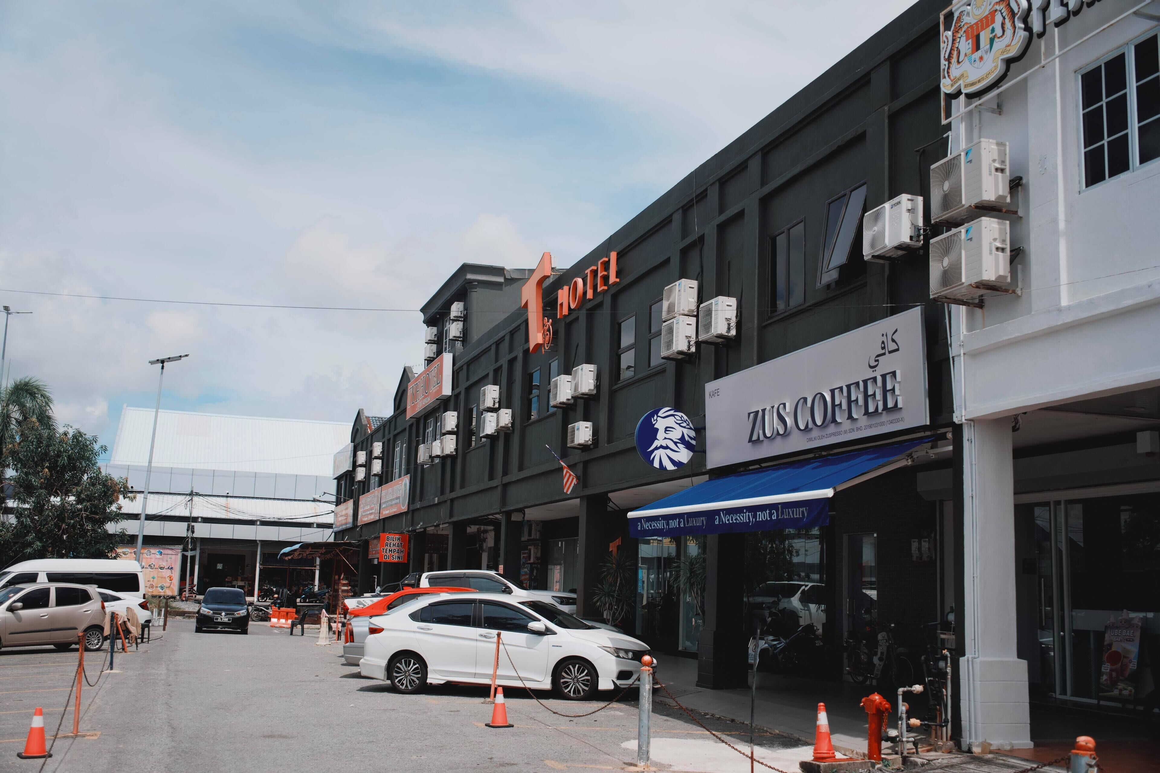 A hotel with a sign reading "HOTEL" and a coffee shop with a sign reading "ZUS COFFEE". There are cars parked in front and traffic cones on the street.