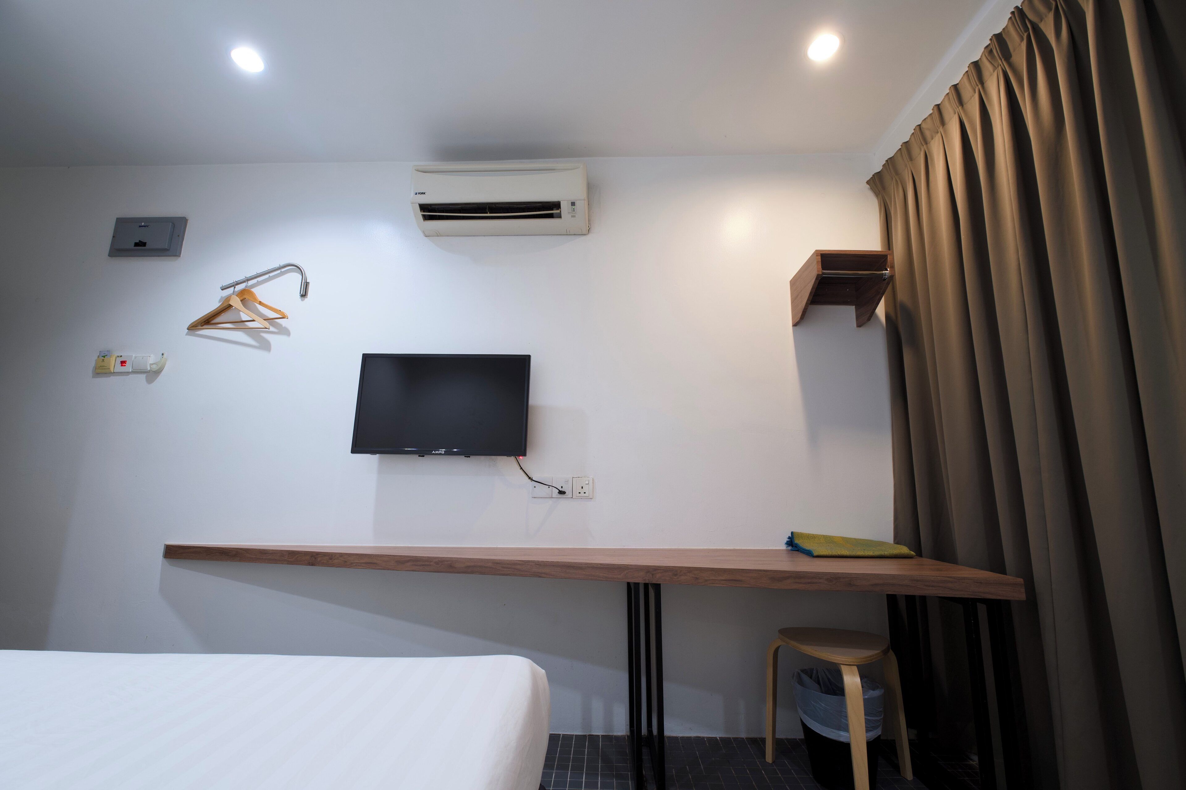 A hotel room with a bed, a desk with a chair, a TV mounted on the wall, an air conditioning unit, a power outlet, and a curtain.