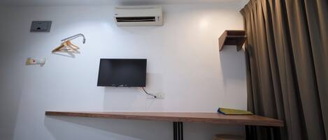 A hotel room with a bed, a desk with a chair, a TV mounted on the wall, an air conditioning unit, a power outlet, and a curtain.