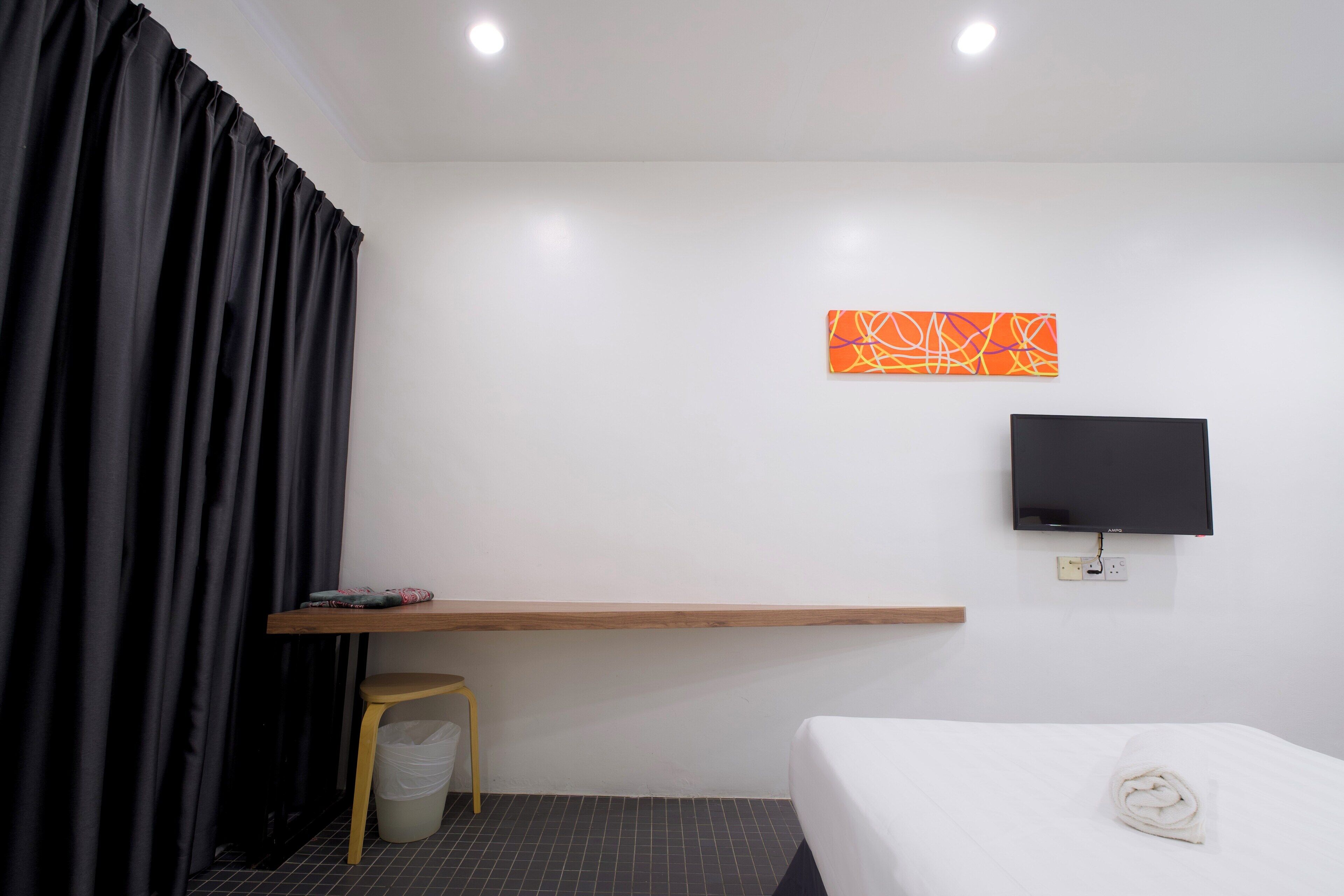 A hotel room with a bed, a TV mounted on the wall, a small wooden desk, a trash bin, and a black curtain.