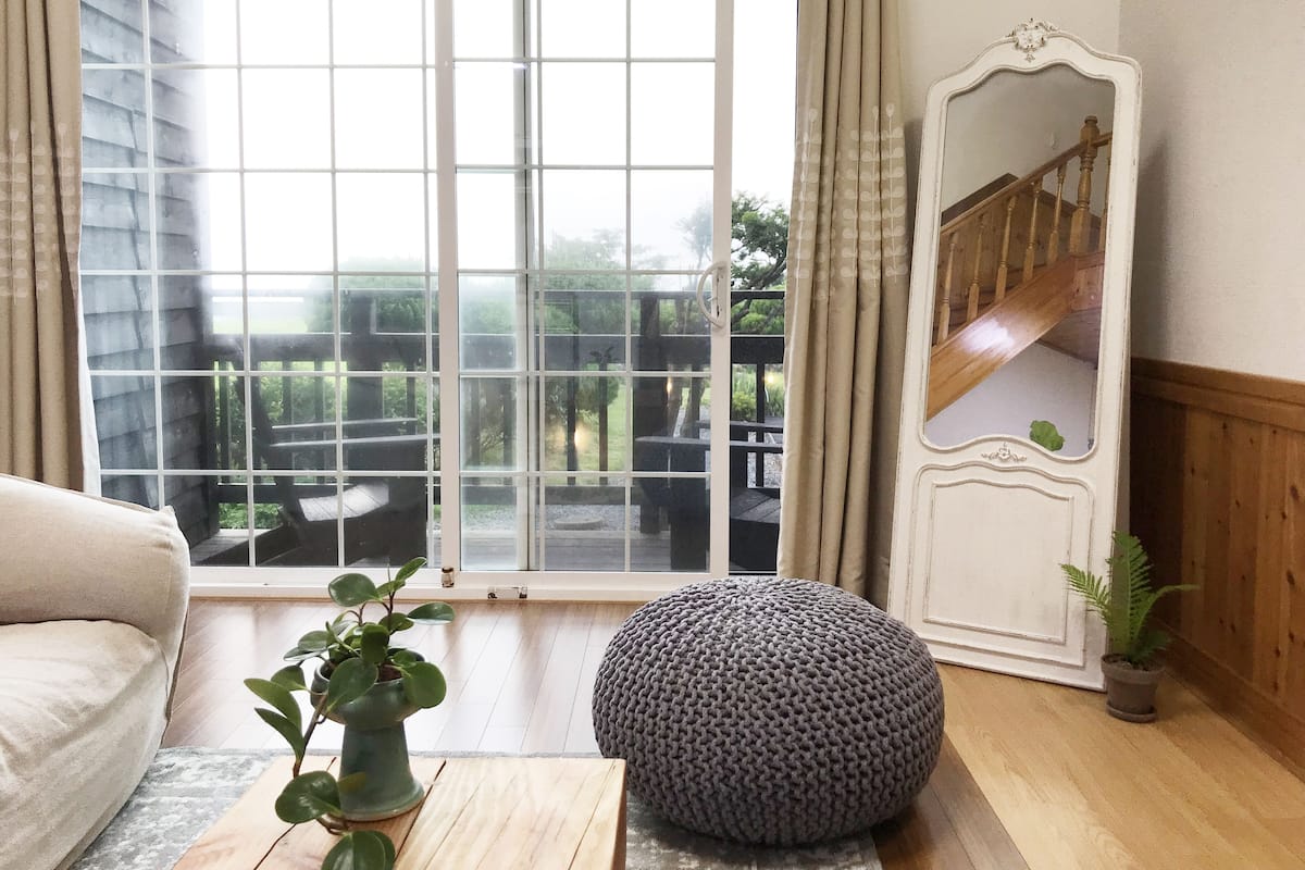 A living room with a large window, a sofa, a round ottoman, a mirror, and a staircase.