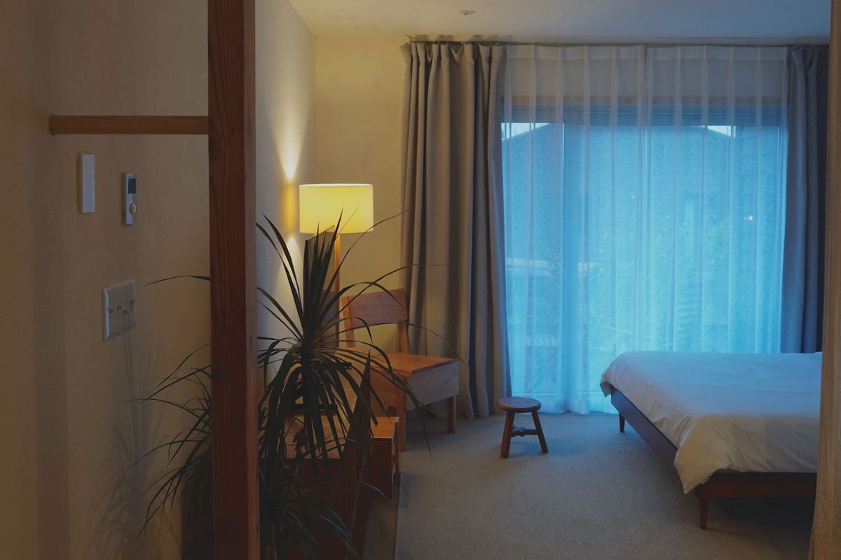 A hotel room with a bed, a desk with a lamp, a potted plant, and a view through a large window.