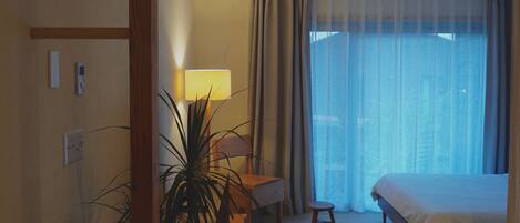 A hotel room with a bed, a desk with a lamp, a potted plant, and a view through a large window.