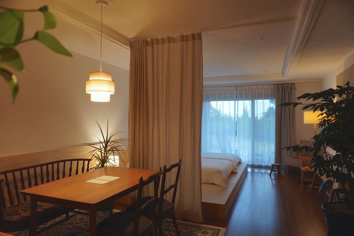 A hotel room with a wooden dining table, chairs, a bed with white bedding, a large window with curtains, and a potted plant.