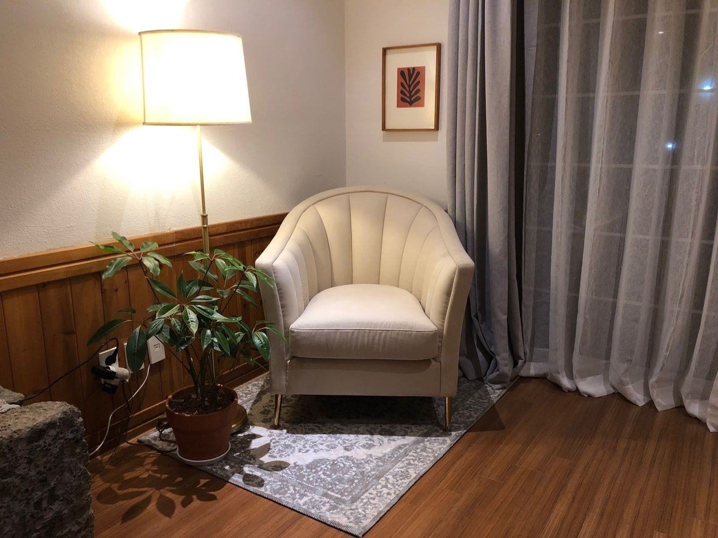 Ein Zimmer mit einem beigefarbenen Sessel, einer Stehlampe, einem Blumentopf, einem gemusterten Teppich und einem gerahmten Bild an der Wand.