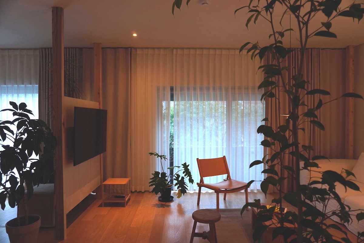 A living room with a flat-screen TV mounted on the wall, a wooden chair, a small stool, and a potted plant.