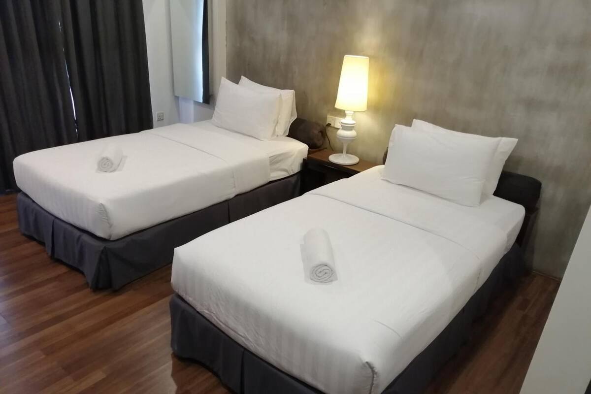 Dos camas individuales con sábanas blancas y faldas oscuras, una mesita de noche con lámpara y una ventana con cortinas.