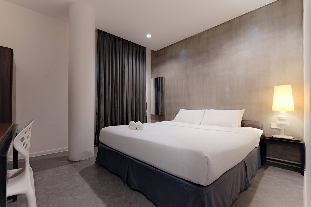 Una habitación de hotel con una cama grande, un escritorio, una silla, una lámpara y un ventanal con cortinas.