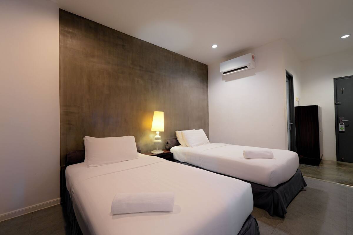 Habitación de hotel con dos camas, aire acondicionado en la pared, lámpara de noche y una puerta a la derecha.