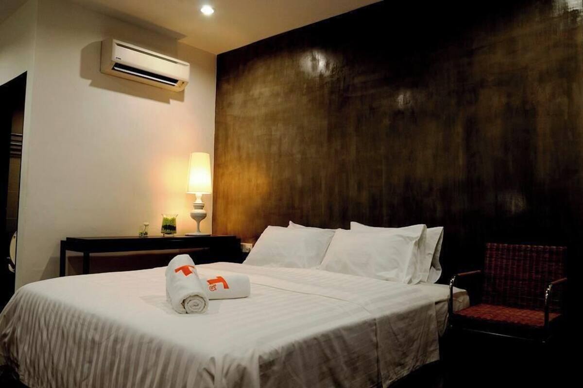 Una habitación de hotel con una cama, una silla, una mesita de noche, una lámpara y un equipo de aire acondicionado.