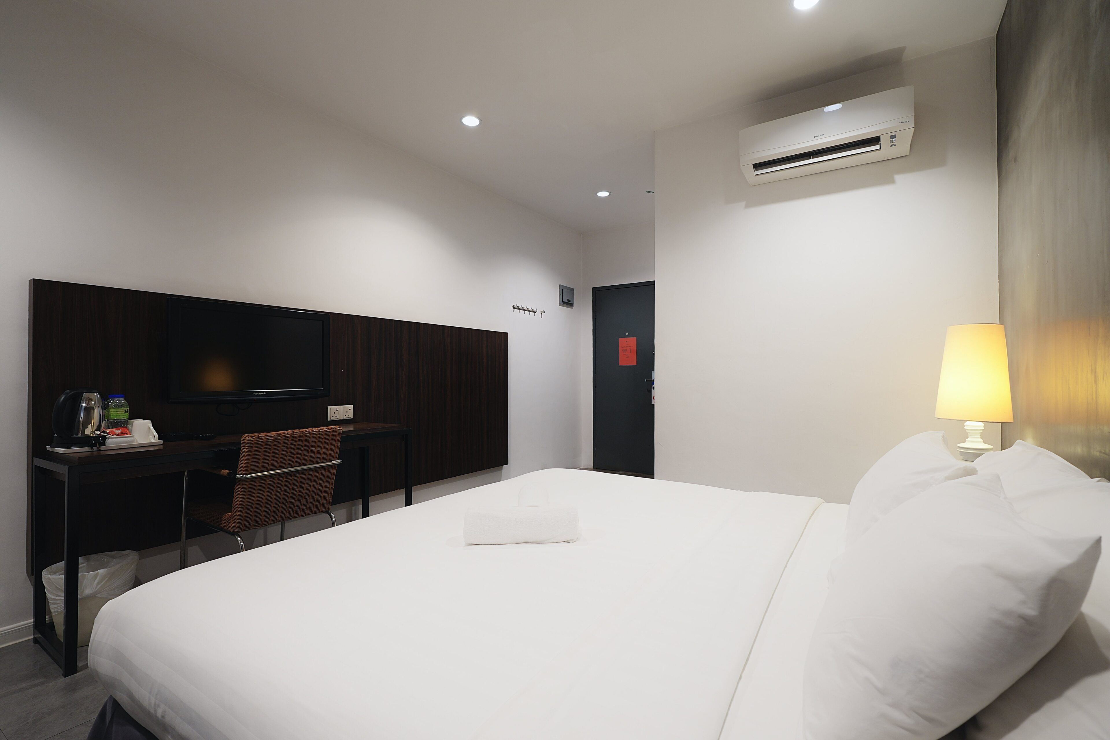 A hotel room with a bed, a desk with a chair, a television, and an air conditioning unit.