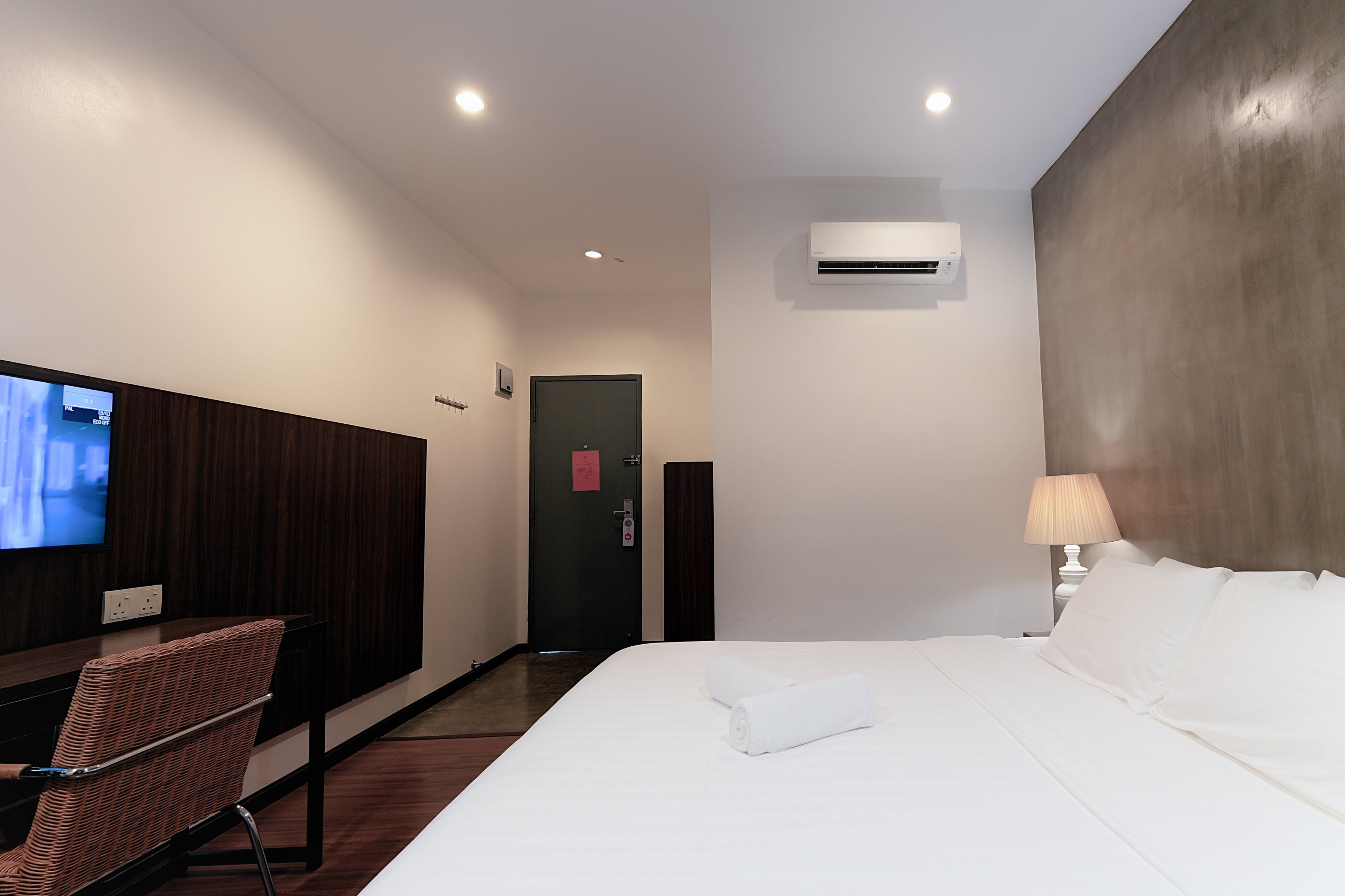 A hotel room with a bed, a TV mounted on the wall, a desk with a chair, and an air conditioning unit.