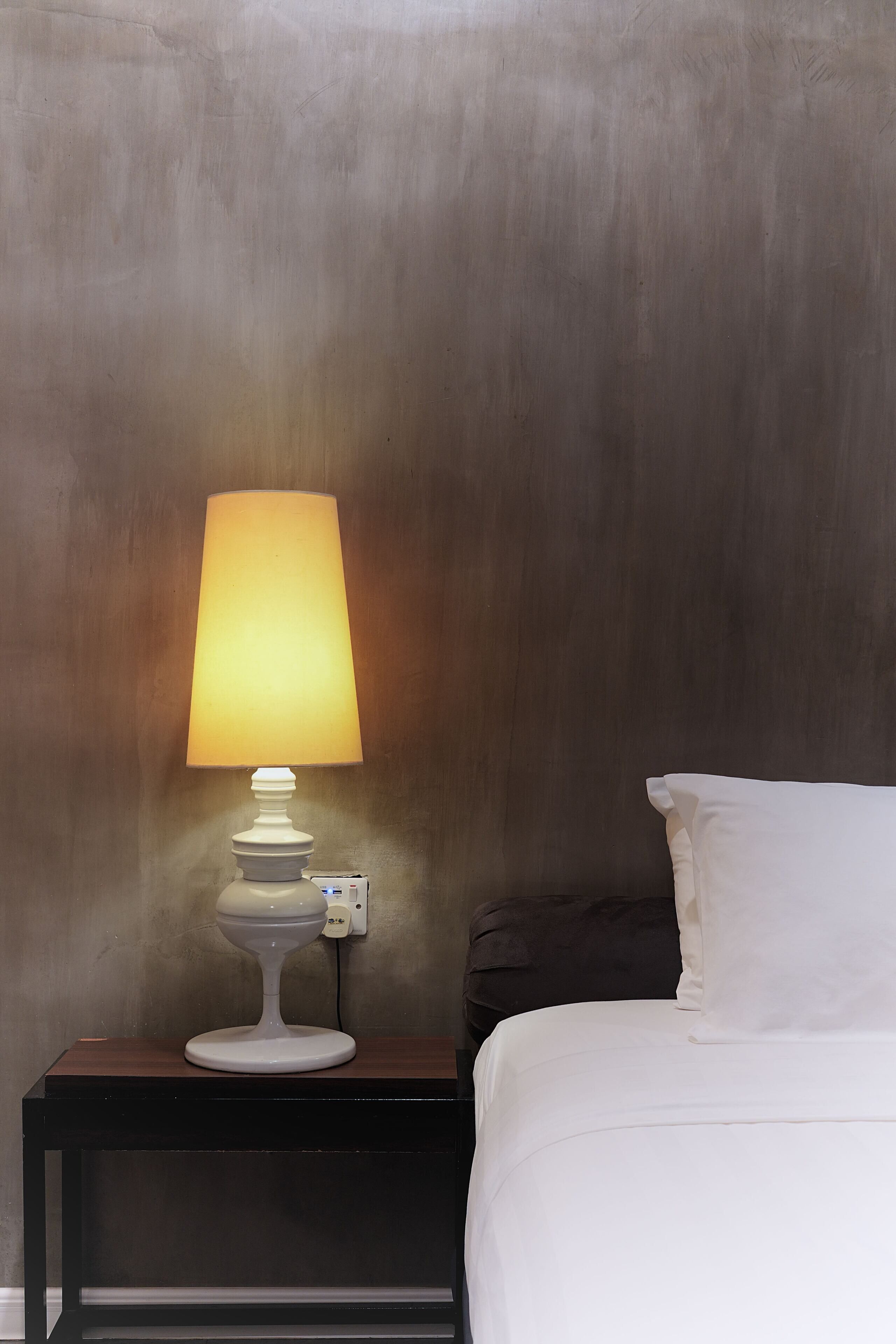 A bed with white bedding, a bedside table with a white lamp, and a dark-colored wall.