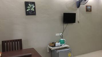 Ein kleines Zimmer mit einem Holztisch, einem grauen Kühlschrank, einem Wandventilator, einem Flachbildfernseher und einem gerahmten Bild an der Wand.