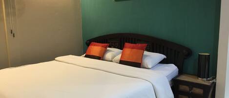 A neatly made bed with white bedding and two orange pillows, a small bedside table with a lamp, and a framed picture on the wall.