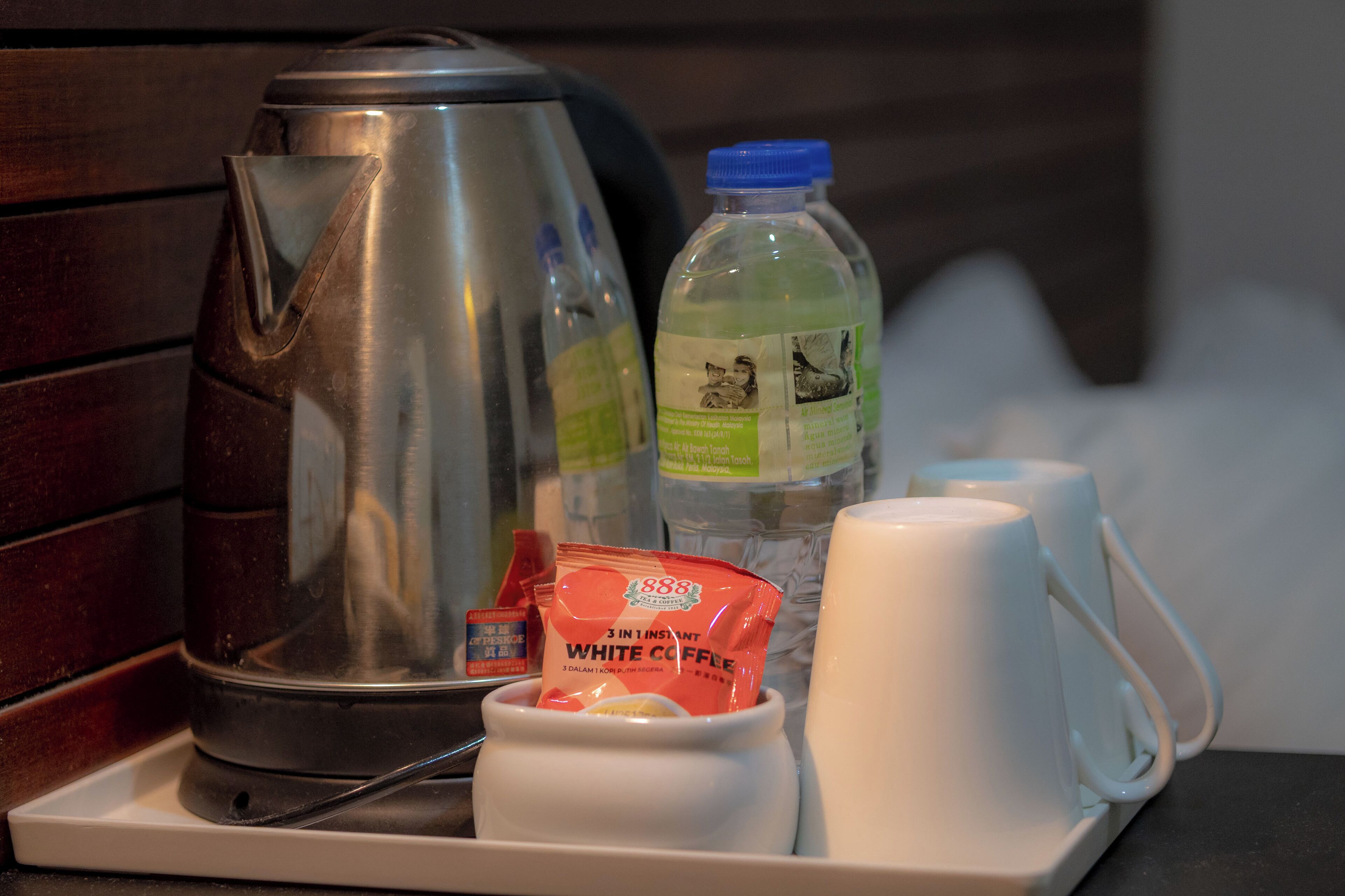 A tray with a stainless steel kettle, a bottle of water, a packet of white coffee, and two white mugs.